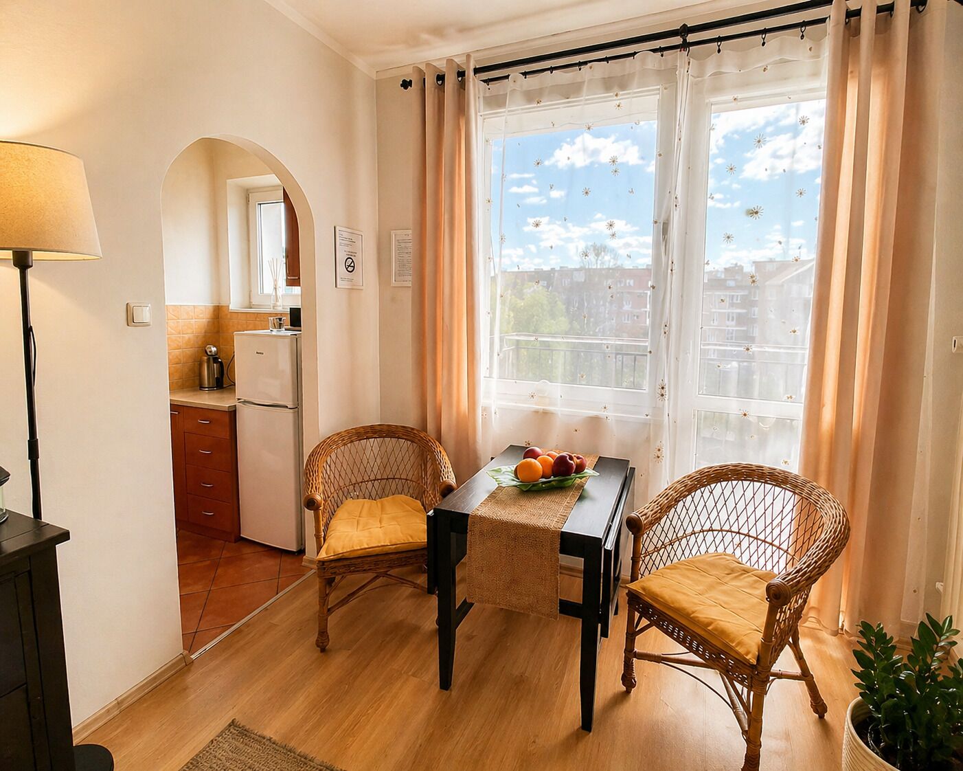 A cozy room with a dining table, two wicker chairs, a small kitchen area, and a window with curtains.