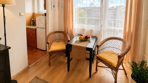 Een gezellige kamer met een eettafel, twee rieten stoelen, een kitchenette en een raam met gordijnen.