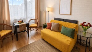 Een kamer met een gele bank, een rieten stoel, een kleine tafel met fruit, een lamp en een lijst met een afbeelding aan de muur.
