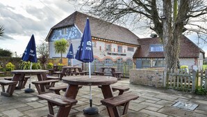 Ein Sitzbereich im Freien mit Holztischen und -bänken, ein Backsteinhaus mit der Aufschrift "The Coach & Horses" und ein Baum im Hintergrund.