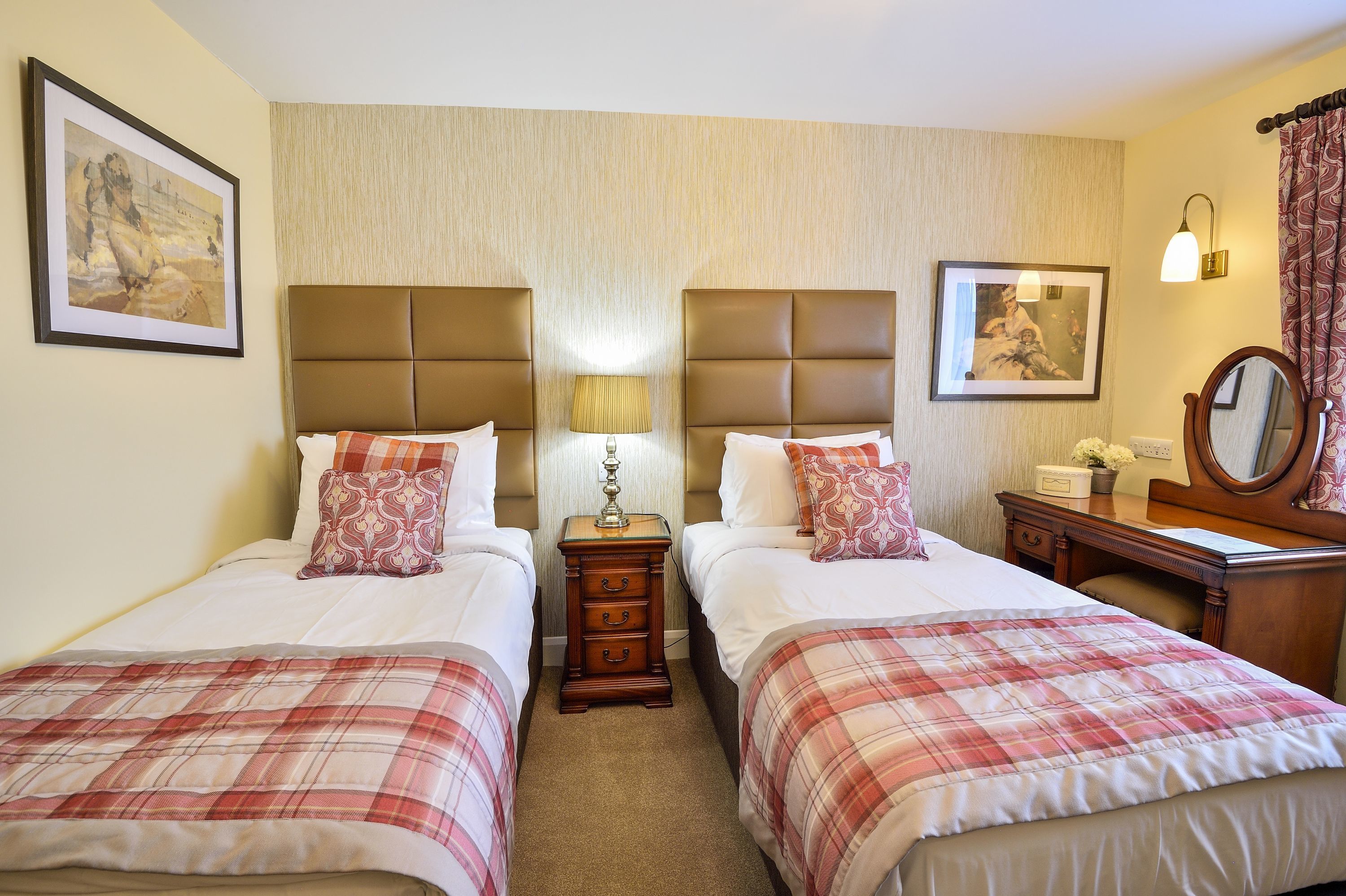 Habitación de hotel con dos camas, una mesita de noche con lámpara, un aparador con espejo y cuadros en las paredes.