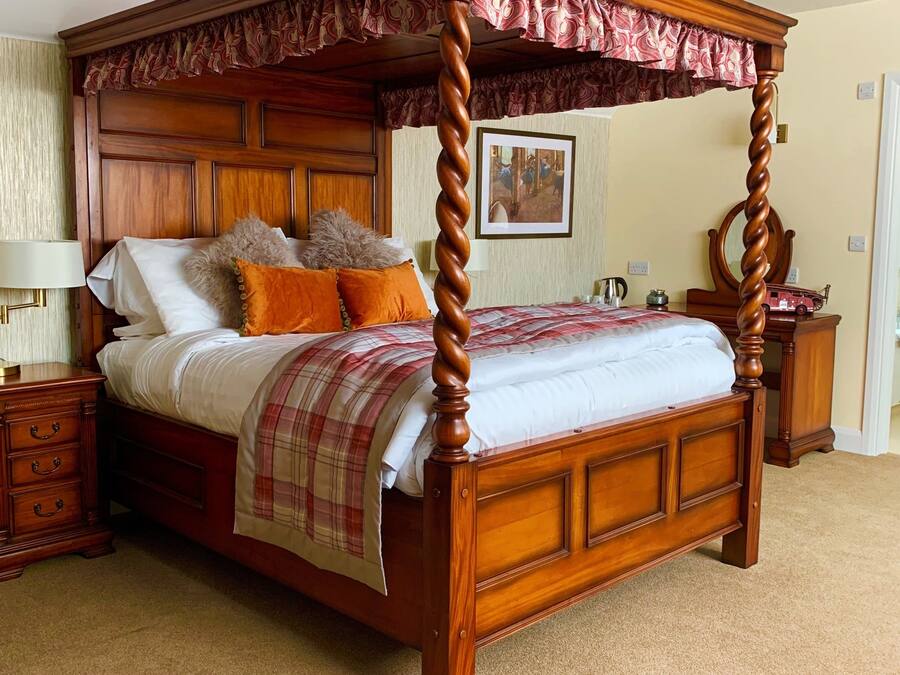 Una cama con dosel de madera y un cubrelecho rojo, una mesita de noche con lámpara, y un cuadro enmarcado en la pared.