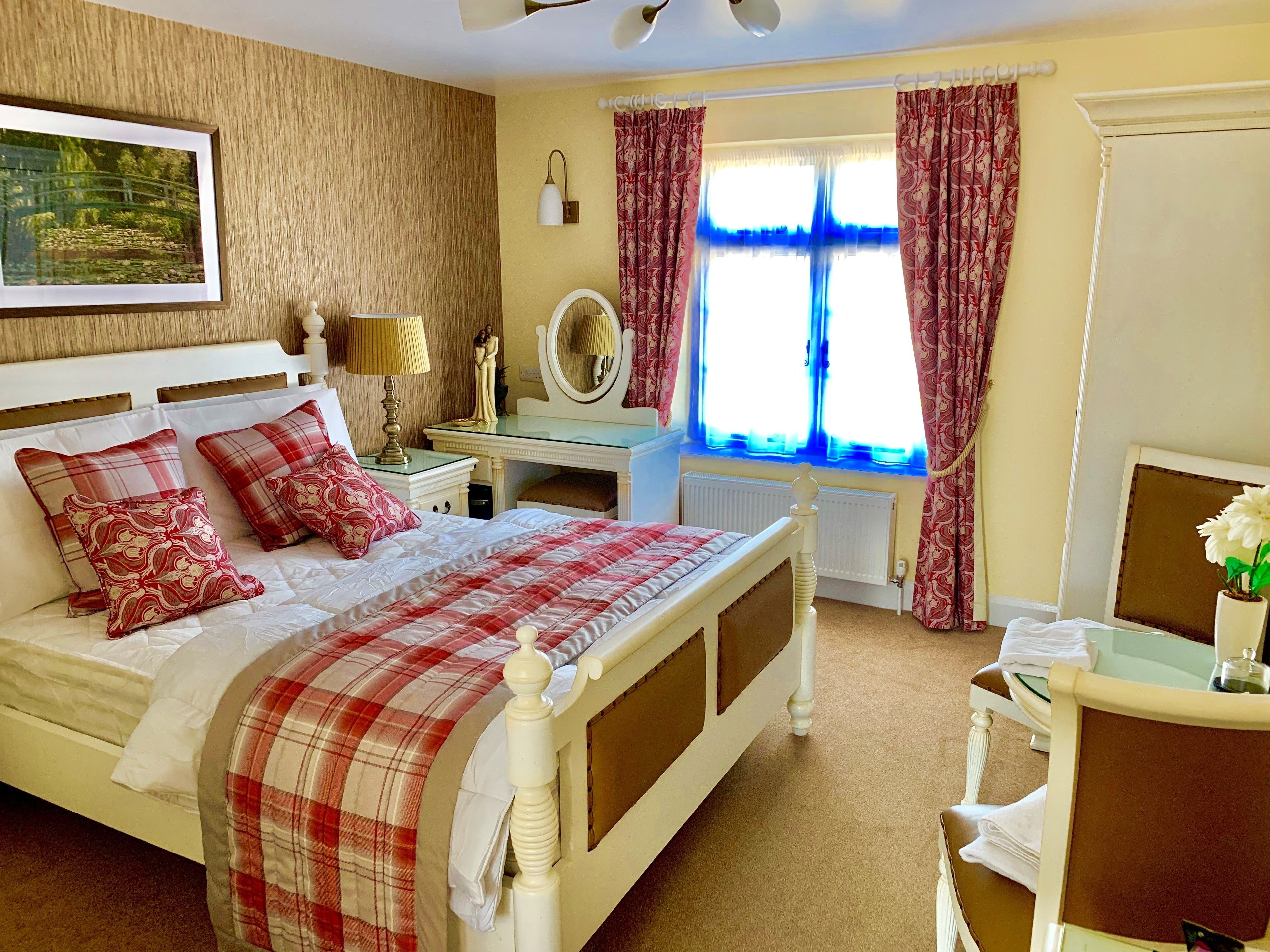 A bedroom with a bed, bedside table, lamp, dresser, window with curtains, and a painting on the wall.