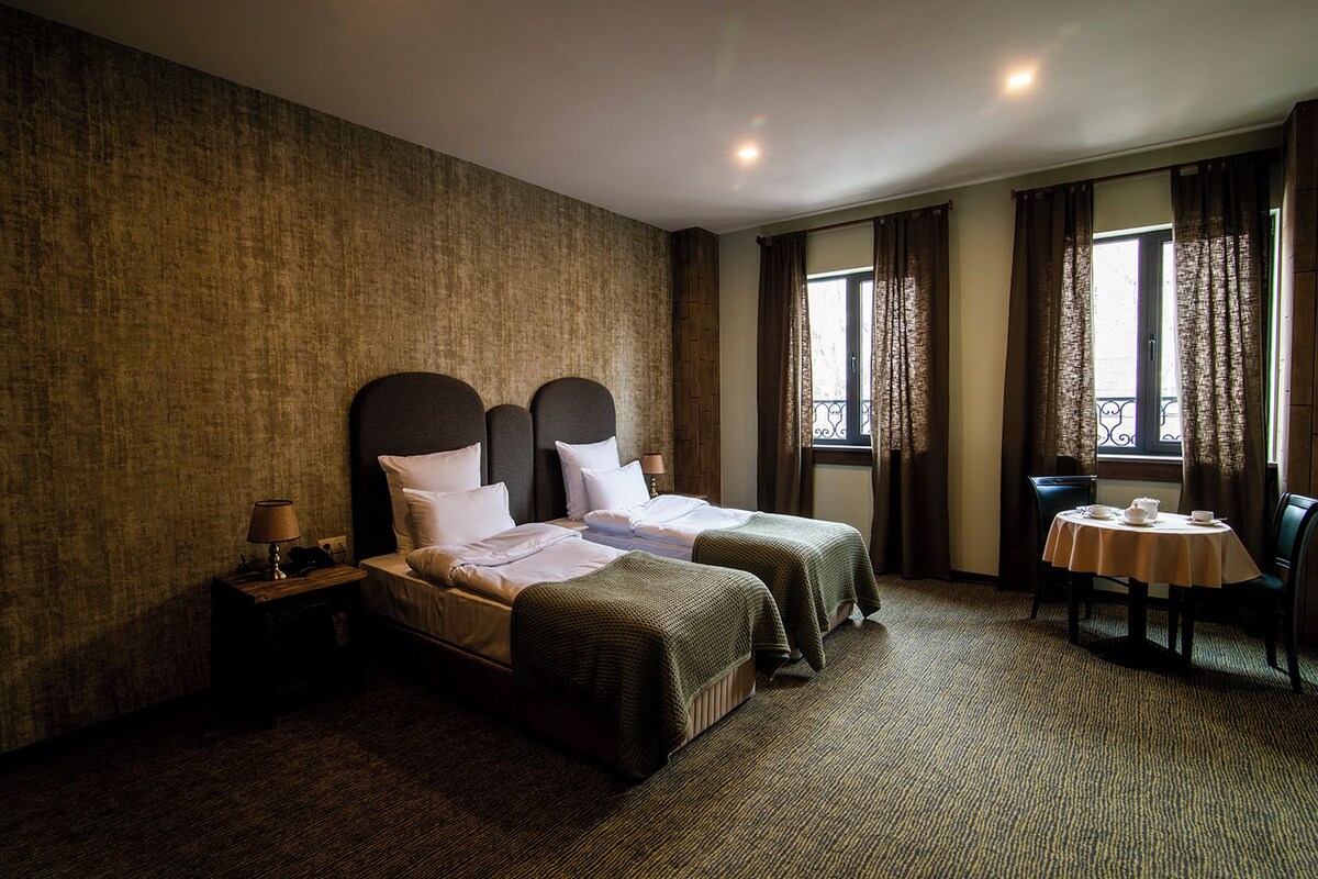 A hotel room with two beds, a small table with a chair, a lamp, and a window with curtains.