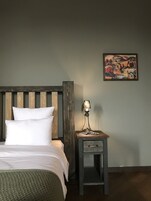 Een bed met witte lakens, een houten hoofdbord, een nachtkastje met een lamp en een lijst met een schilderij aan de muur.