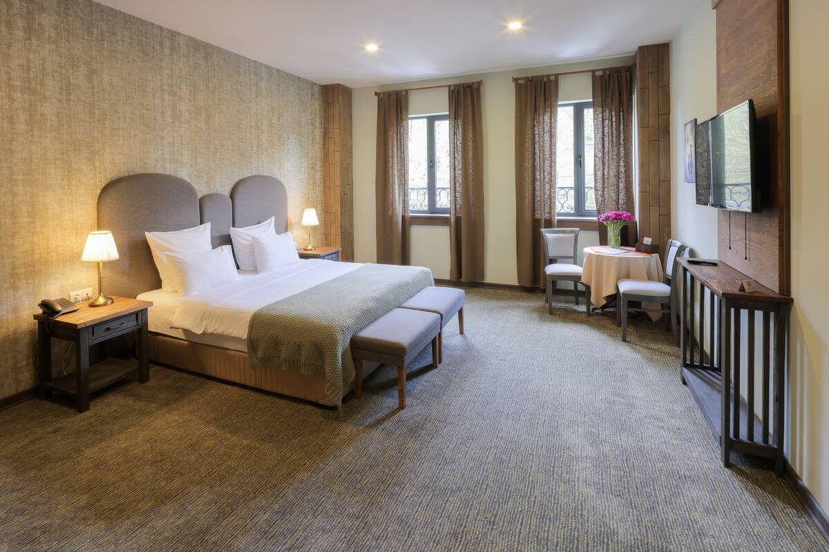 A hotel room with a large bed, bedside tables, a desk with a chair, a TV mounted on the wall, and a window with curtains.