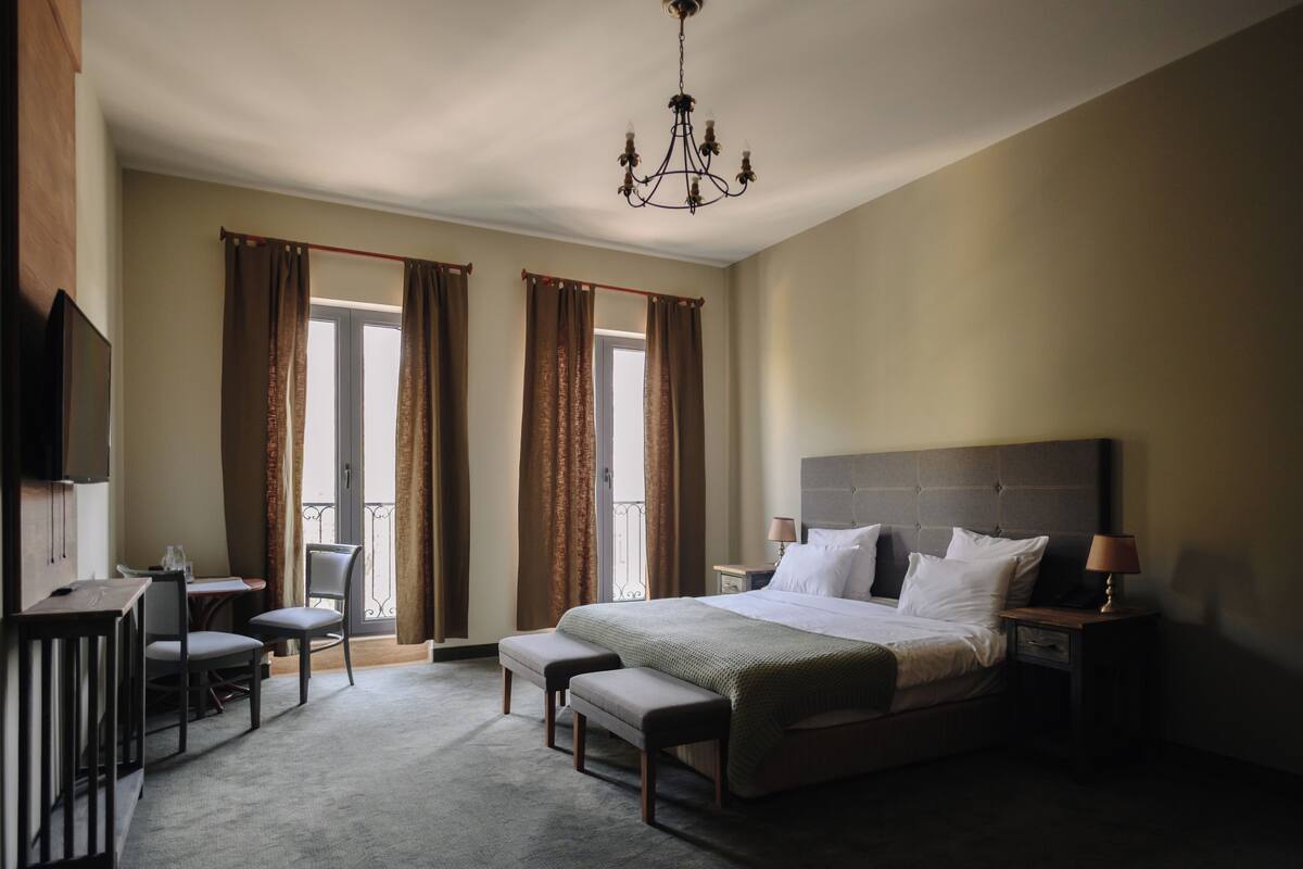 A hotel room with a large bed, a desk with a chair, a bench, a TV, and a chandelier.