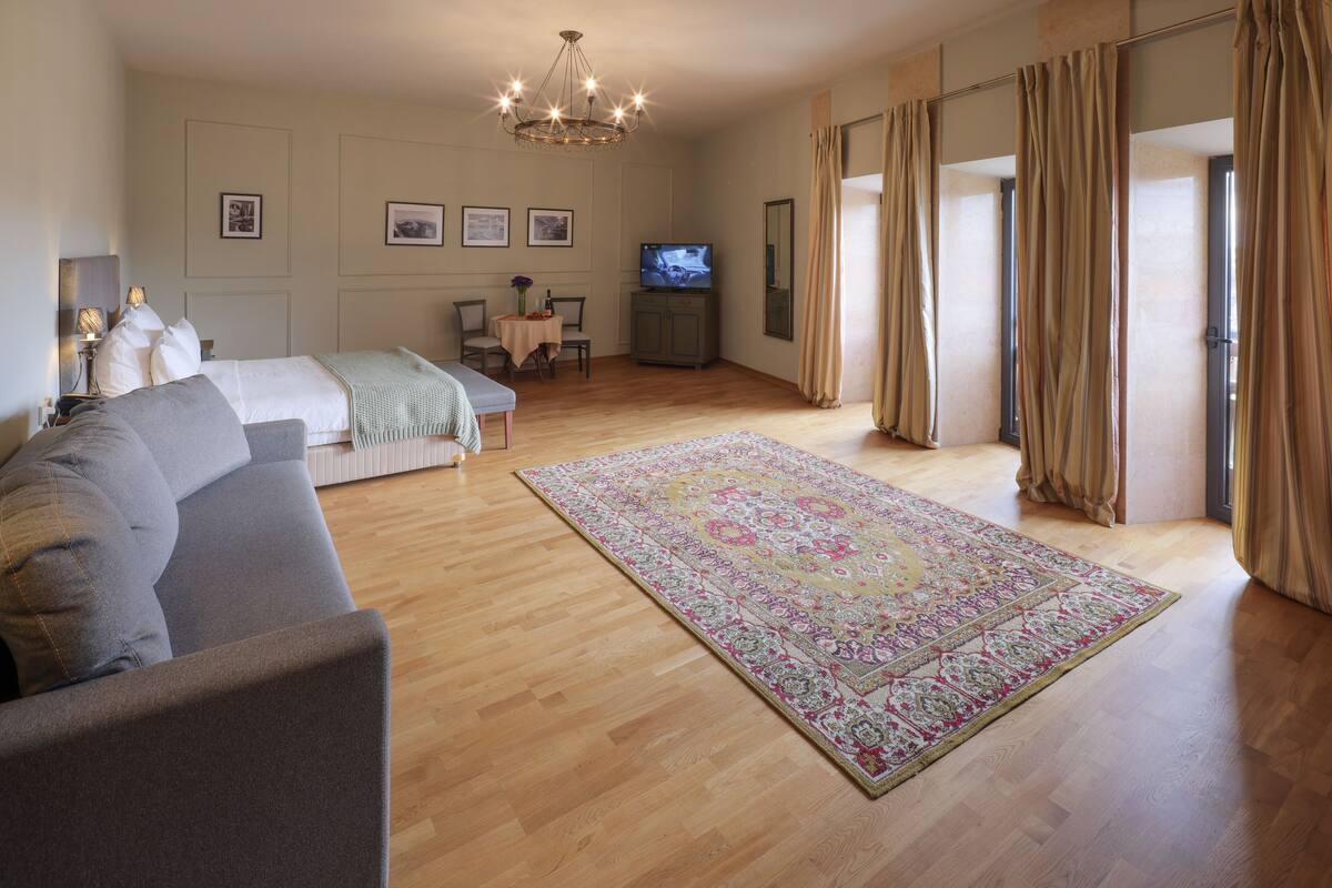 A spacious hotel room with a large bed, a sofa, a television, a dining table with chairs, and a large window with curtains.