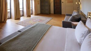Habitación de hotel con una cama grande, un sofá, una araña y piso de madera, con vistas al exterior.