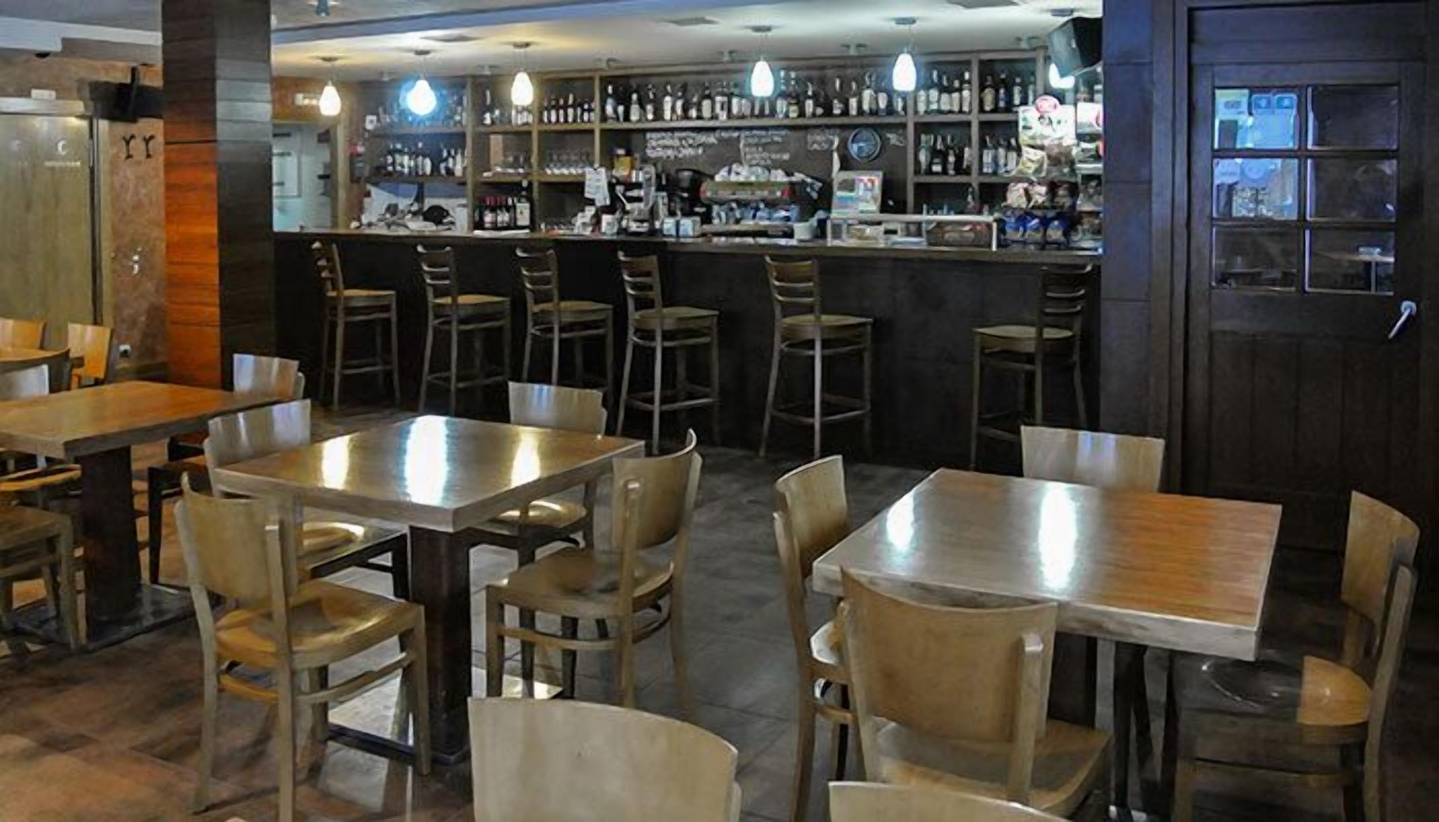 Un restaurante con varias mesas y sillas, una zona de bar con estantes llenos de botellas, y una puerta de madera.