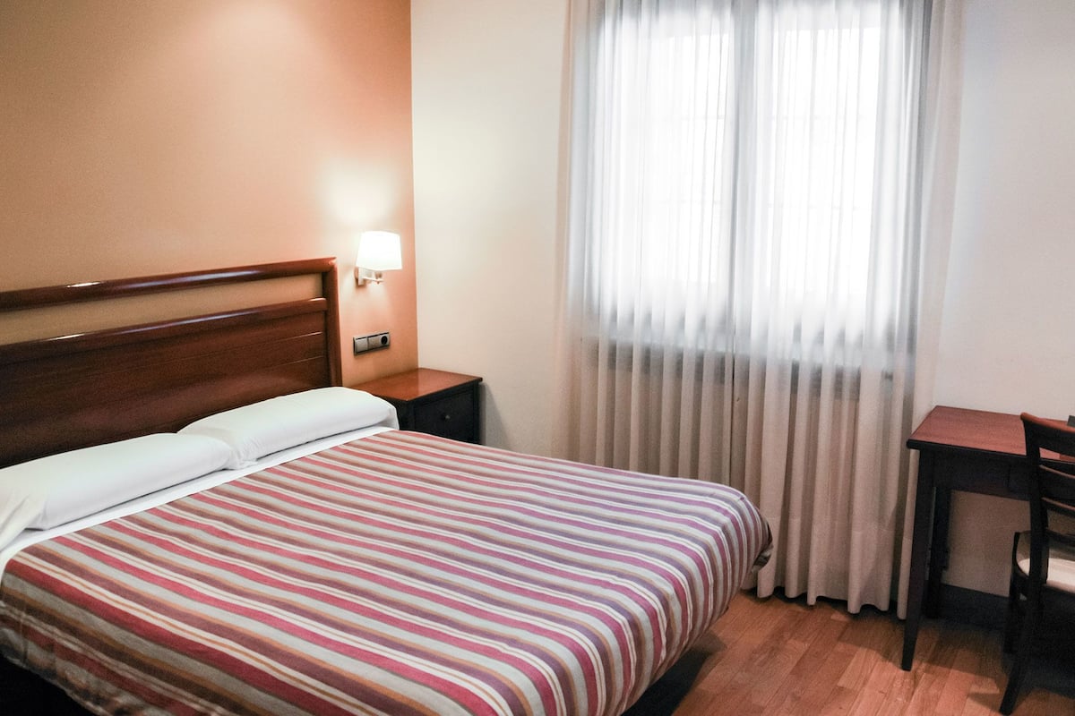 Una habitación de hotel con cama, mesita de noche, escritorio y ventana con cortinas.