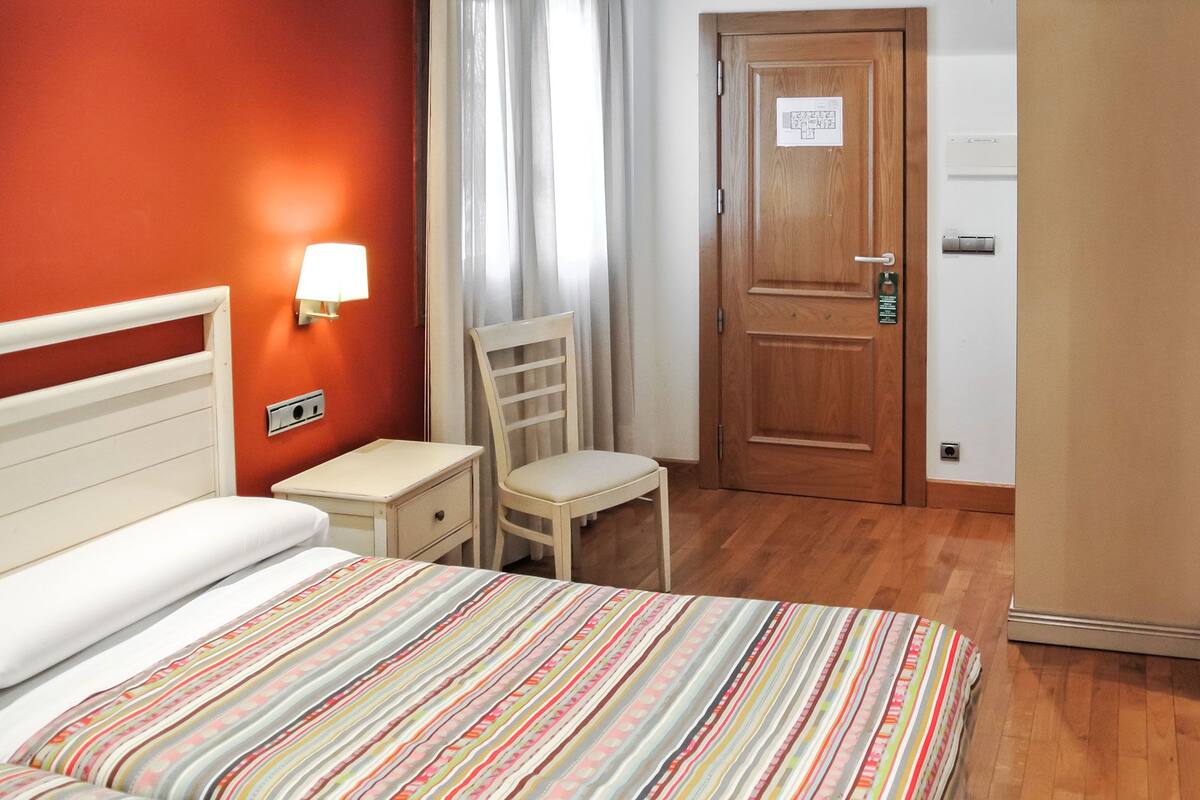Un dormitorio con cama, mesita de noche, silla y una puerta con un cartel.