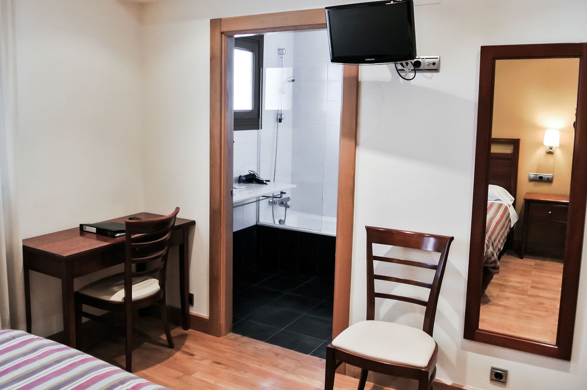 Una habitación de hotel pequeña con escritorio, dos sillas, cama, televisor, espejo y un baño visible a través de una puerta abierta.