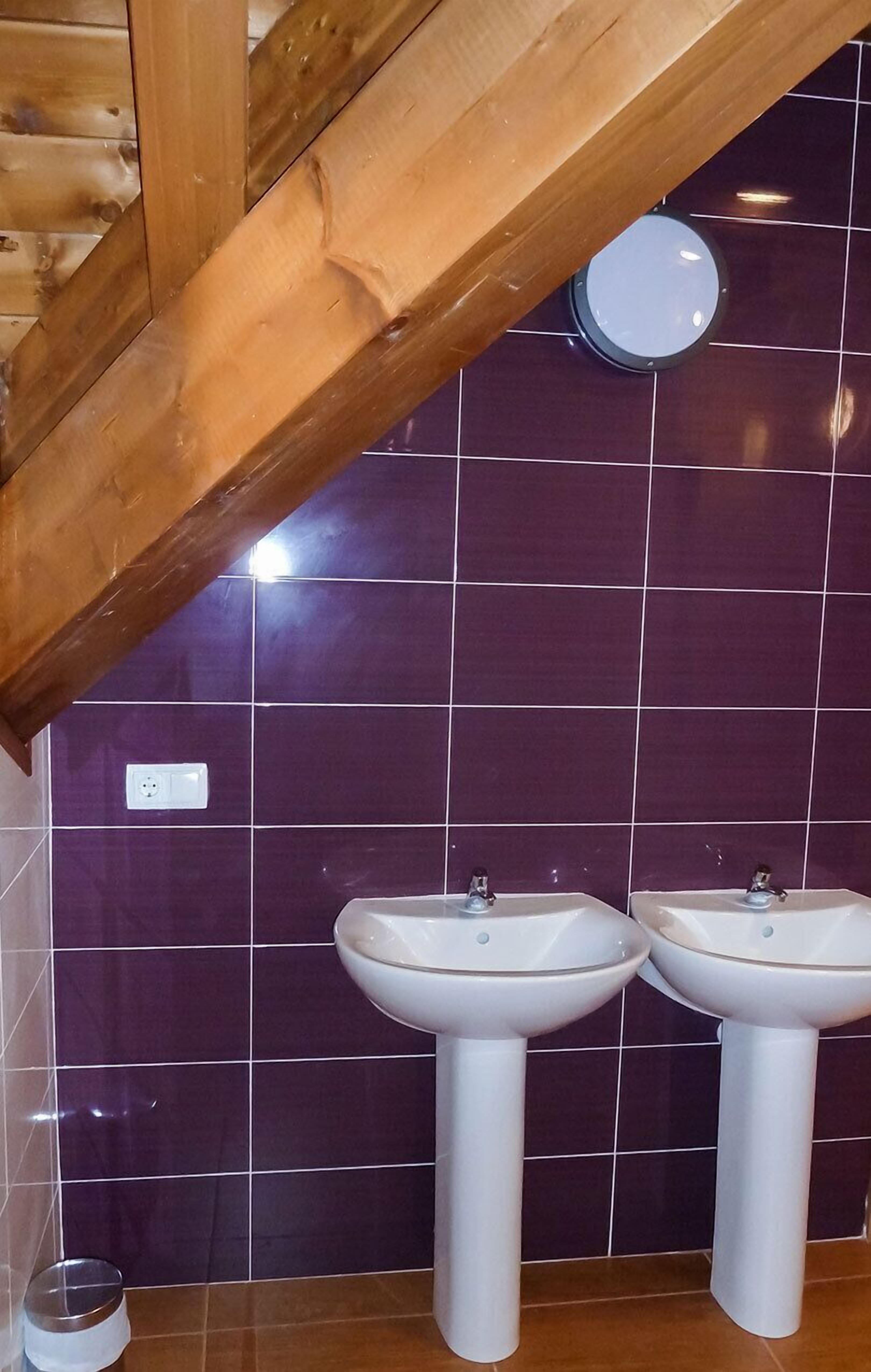 Een badkamer met paars betegelde wanden, twee witte wastafels op voetstukken, en een houten balk in het plafond.