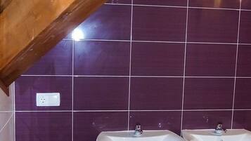 Een badkamer met paars betegelde wanden, twee witte wastafels op voetstukken, en een houten balk in het plafond.