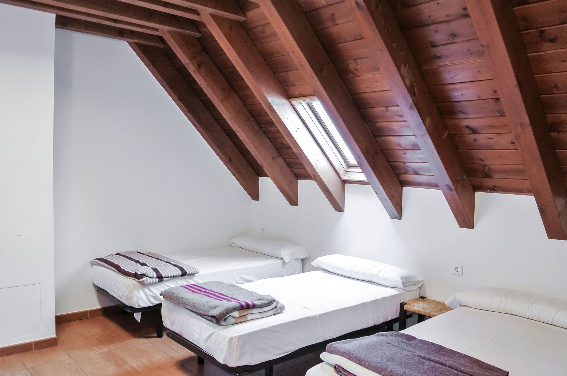 A small, cozy room with two beds, wooden beams on the ceiling, and a skylight.