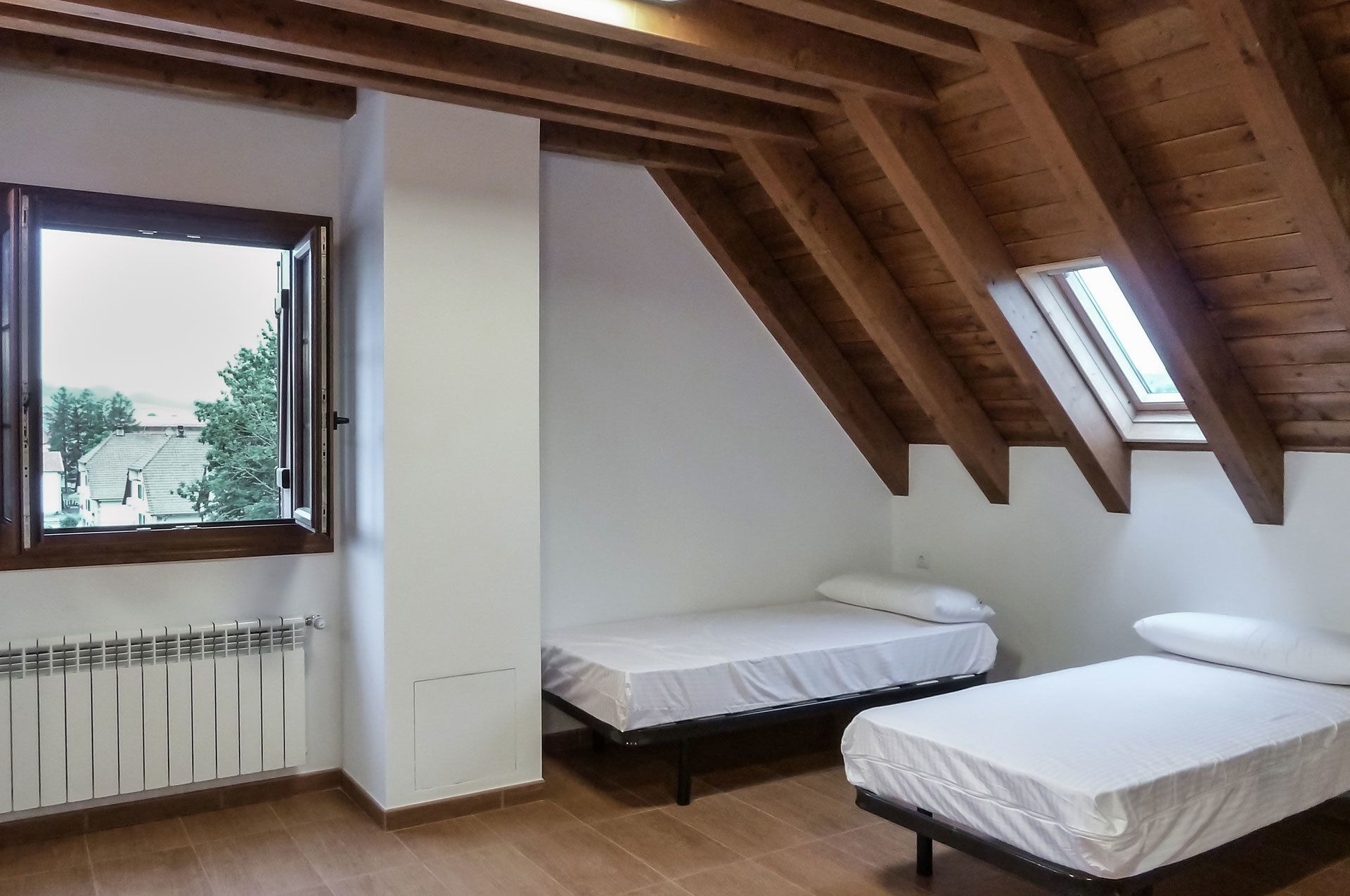 A room with two single beds, a radiator, a window, and a sloped ceiling with wooden beams.