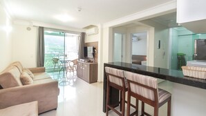 A modern living area with a sofa, a dining table with chairs, a kitchen area with a counter and stools, and a view through a glass door. - Apartamento 1 quarto Summer Beach - 320 (Bombinhas)