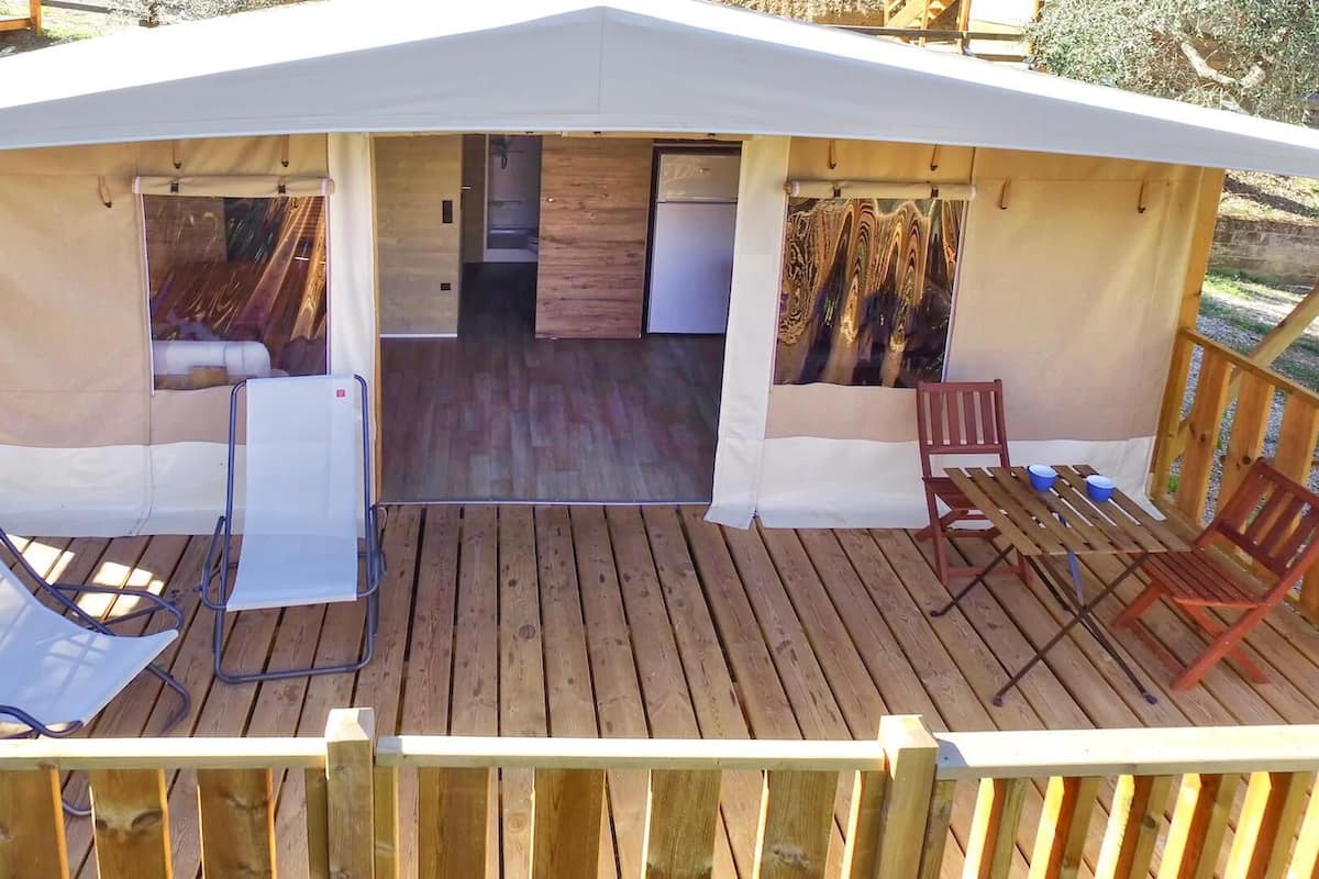 A covered outdoor area with a wooden deck, a table, two chairs, and a small kitchen area with a refrigerator.