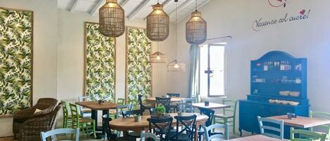 Un café chaleureux avec des tables et des chaises en bois, des lampes en osier suspendues et un comptoir bleu où est affiché le menu.