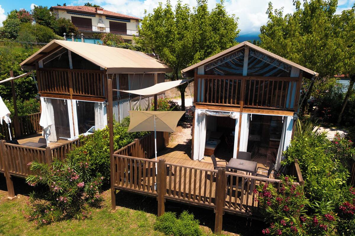 A wooden deck with outdoor seating and a covered area, surrounded by greenery and a view of a building in the background.