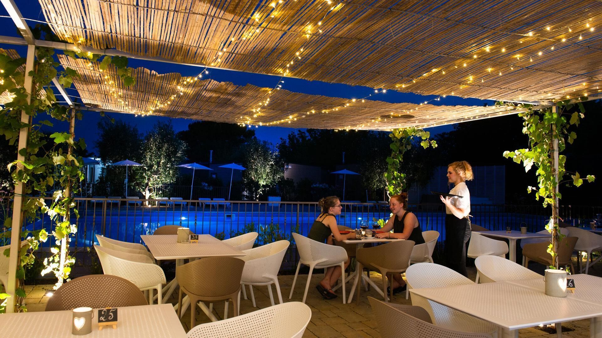 Uma área de refeições ao ar livre com mesas e cadeiras, luzes de fada e uma piscina ao fundo.