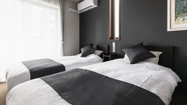 Een hotelkamer met twee bedden, een groot raam met dunne gordijnen, een airconditioner, een nachtkastje en een lijst aan de muur.