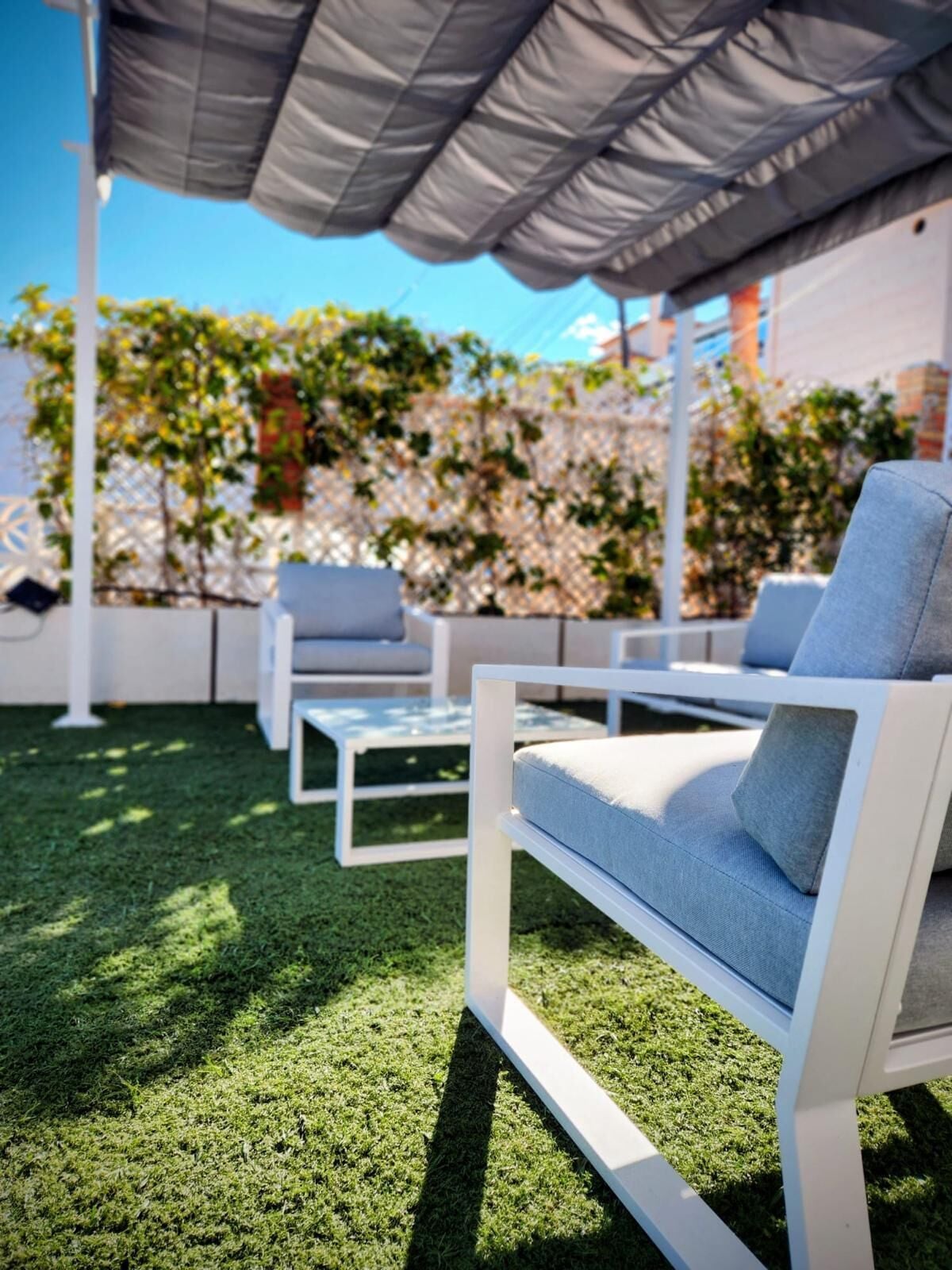 Ein Patio mit weißer Gartenmöblierung, darunter eine Couch und Stühle, unter einem grauen Sonnensegel.