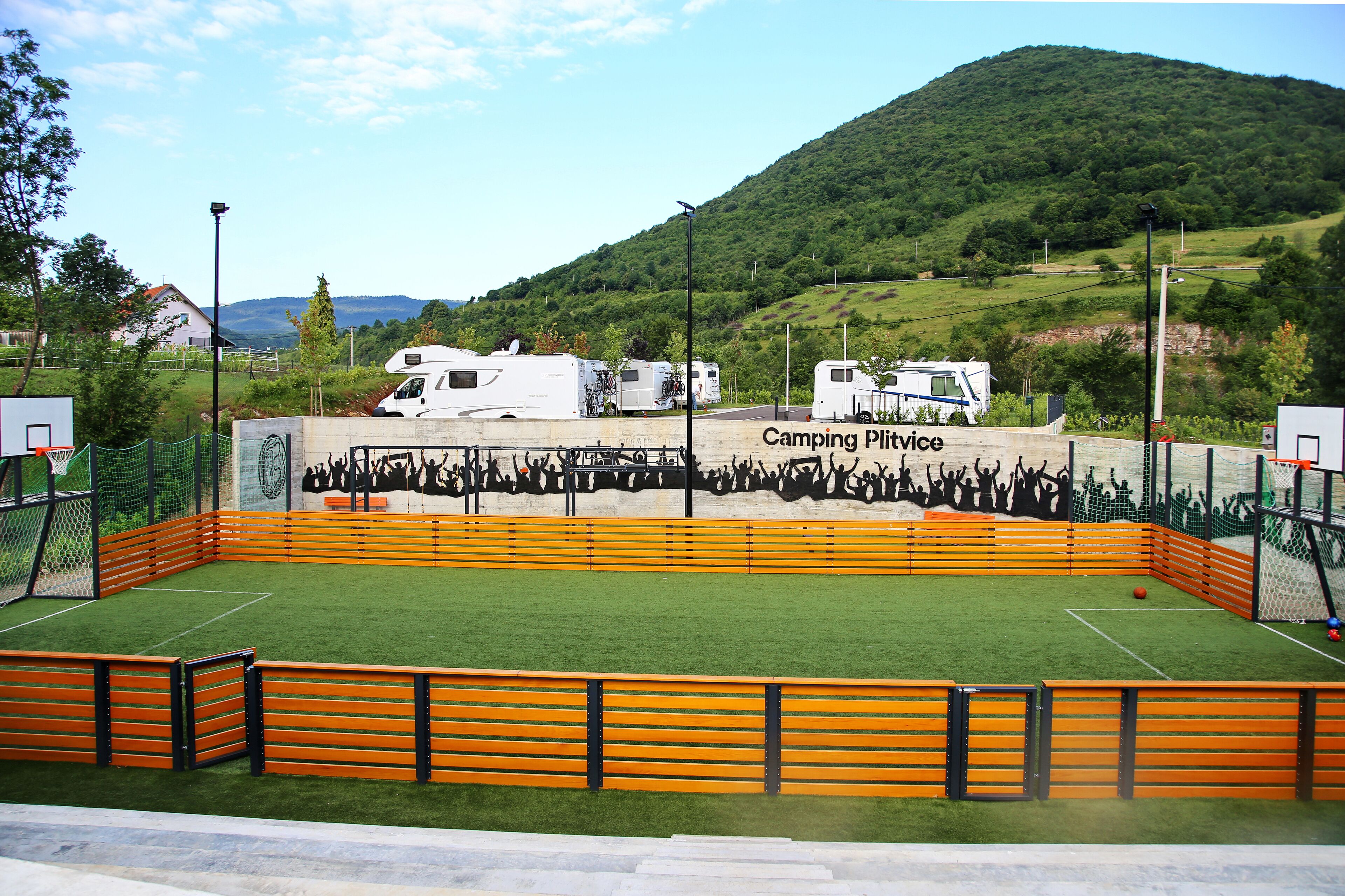 A sports field with a basketball hoop, surrounded by orange fencing, and RVs in the background.