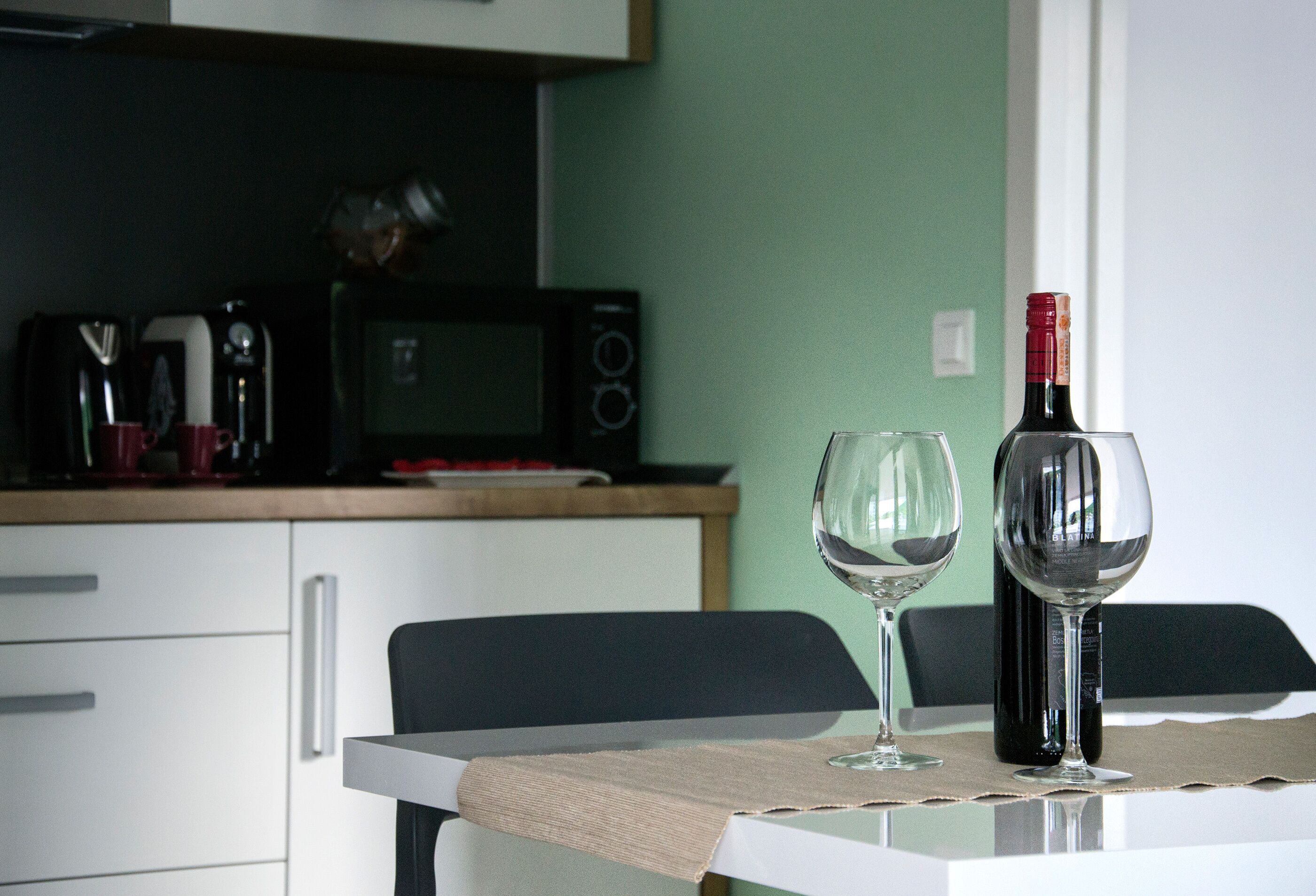 Una mesa con una botella de vino y dos copas, un microondas y una cafetera en la encimera de la cocina.
