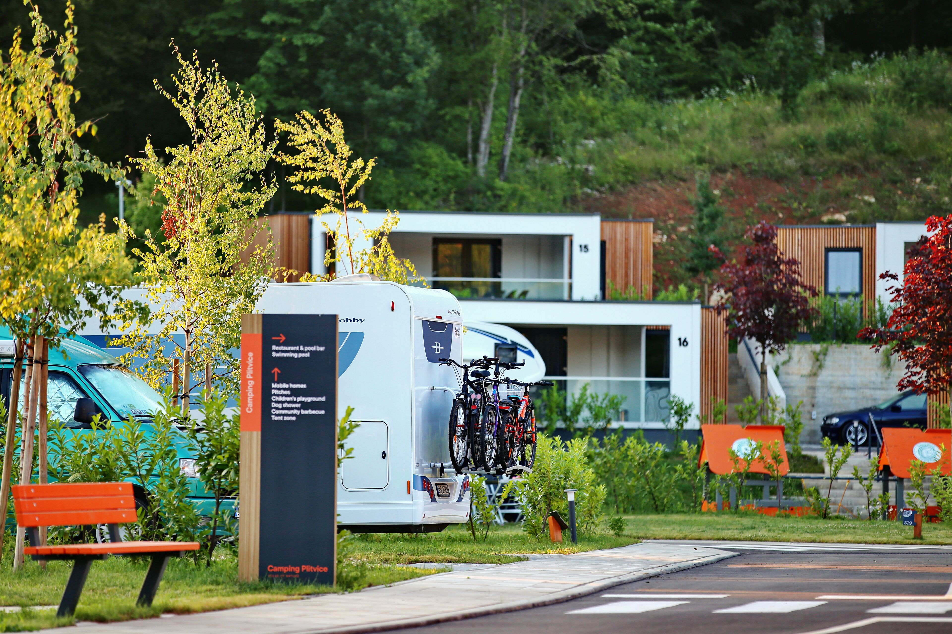 En modern husbil med cyklar ställda på ett bagagehållare baktill, en bänk och en skylt som visar var campingplatsen ligger och vilka faciliteter som finns.