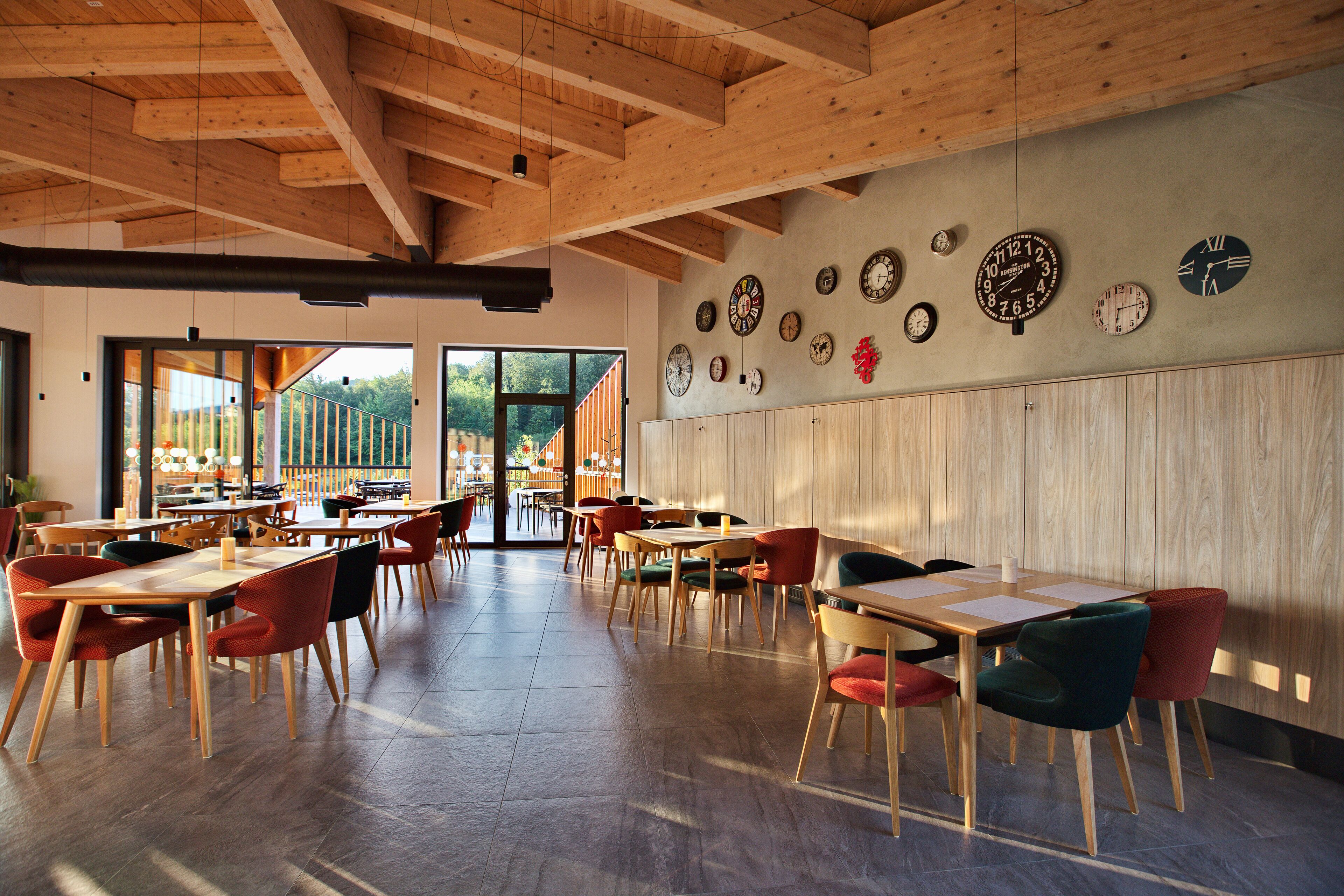 A spacious dining area with wooden tables and chairs, a variety of clocks on the wall, and large windows offering a view of the outdoors.