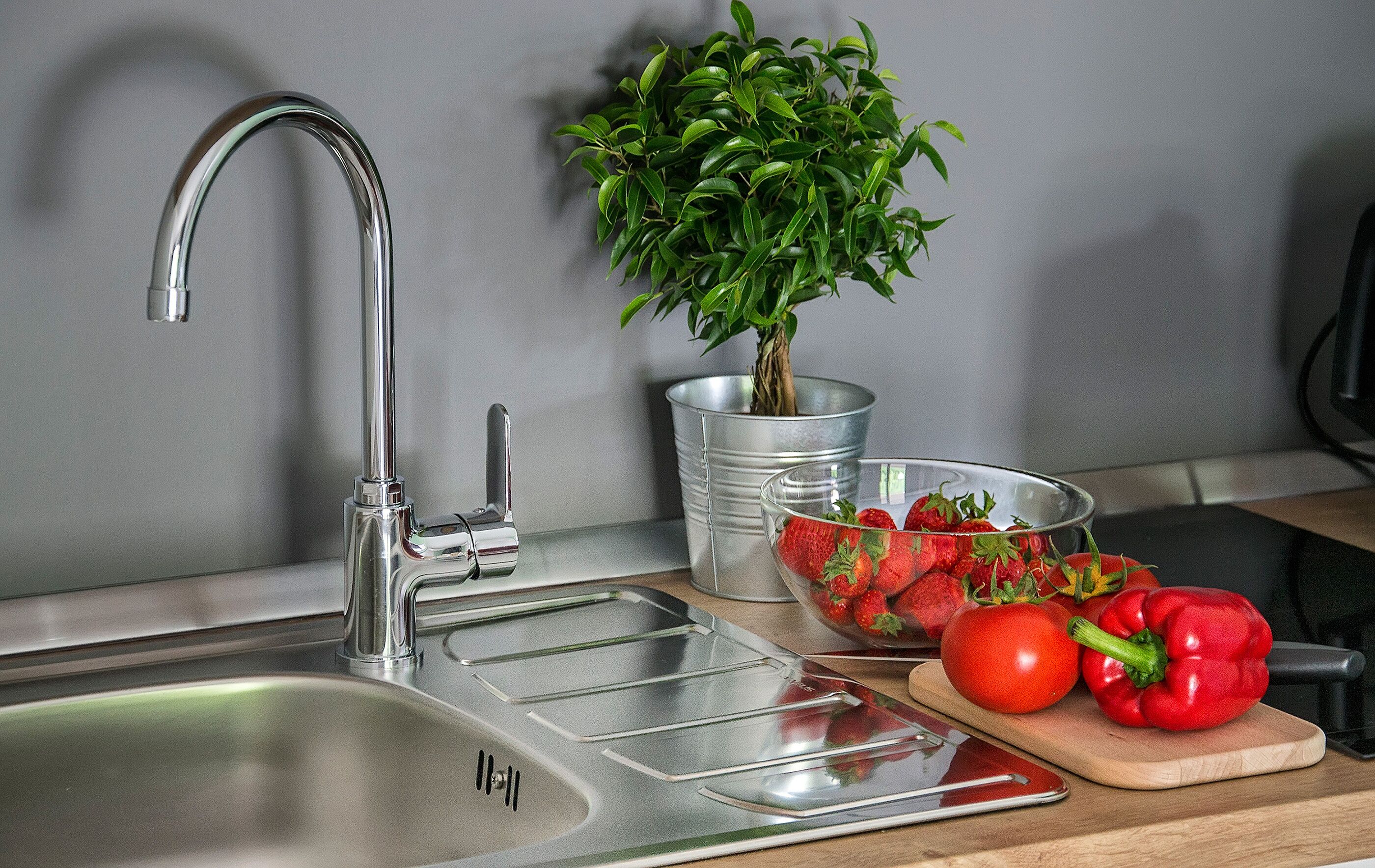 A kitchen sink with a modern faucet, a potted plant, a bowl of strawberries, a tomato, and a red bell pepper on a wooden cutting board.