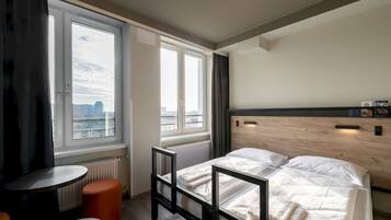 Habitación de hotel moderna con una cama, una pequeña mesa y vistas de la ciudad a través de grandes ventanales.