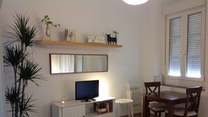 Una sala de estar con televisor, un mueble blanco, una mesa de comedor, sillas y una ventana con persianas.