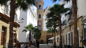 Una calle adoquinada bordeada de palmeras y edificios, con zonas de estar al aire libre y una imponente torre de iglesia al fondo.