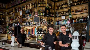 Un bar con estantes llenos de botellas diversas, una barra y dos camareros.