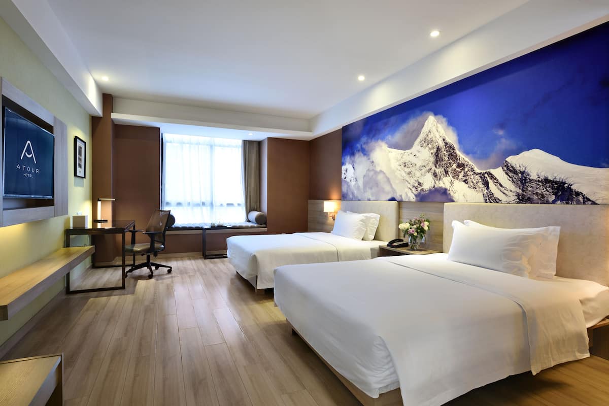 Quarto de hotel com duas camas, uma escrivaninha, uma cadeira, uma televisão e uma grande pintura de montanhas na parede.