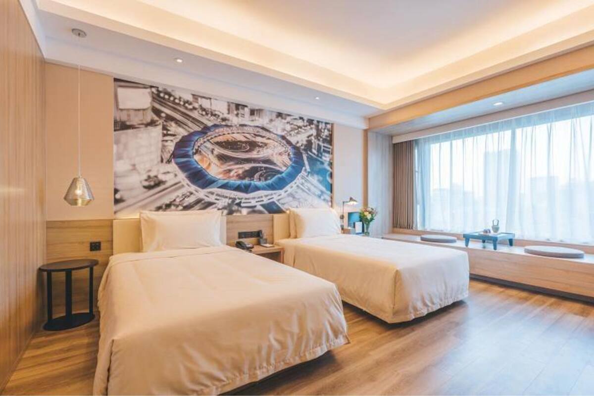 Une chambre d’hôtel avec deux lits, une grande peinture abstraite au mur, une grande fenêtre avec des rideaux et une petite table avec une lampe.