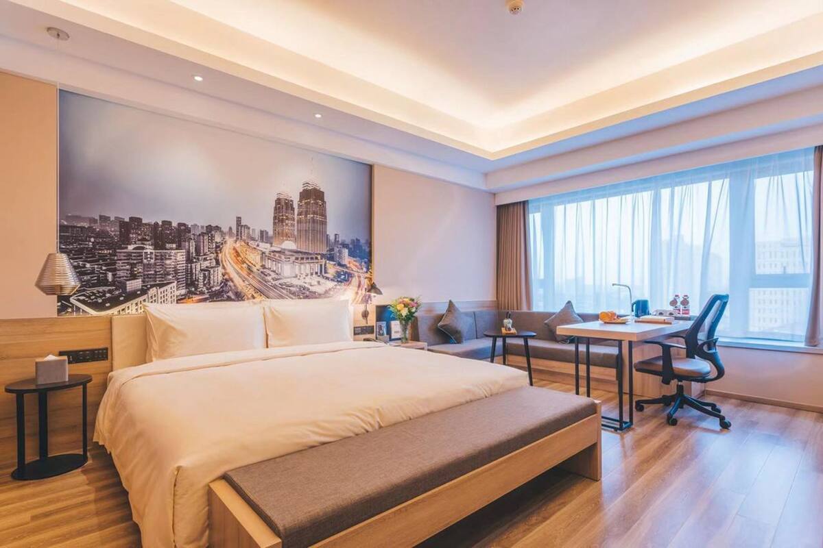 Une chambre d’hôtel avec un grand lit, un bureau et une chaise. Il y a un tableau au mur et une fenêtre avec des rideaux.