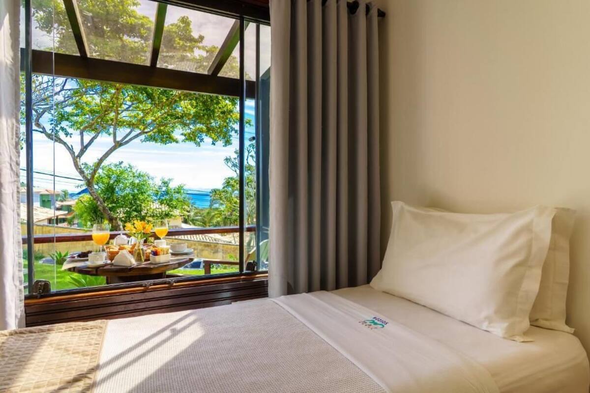 Habitación de hotel con cama, ventana con cortinas y vista a un balcón con mobiliario exterior y un árbol.