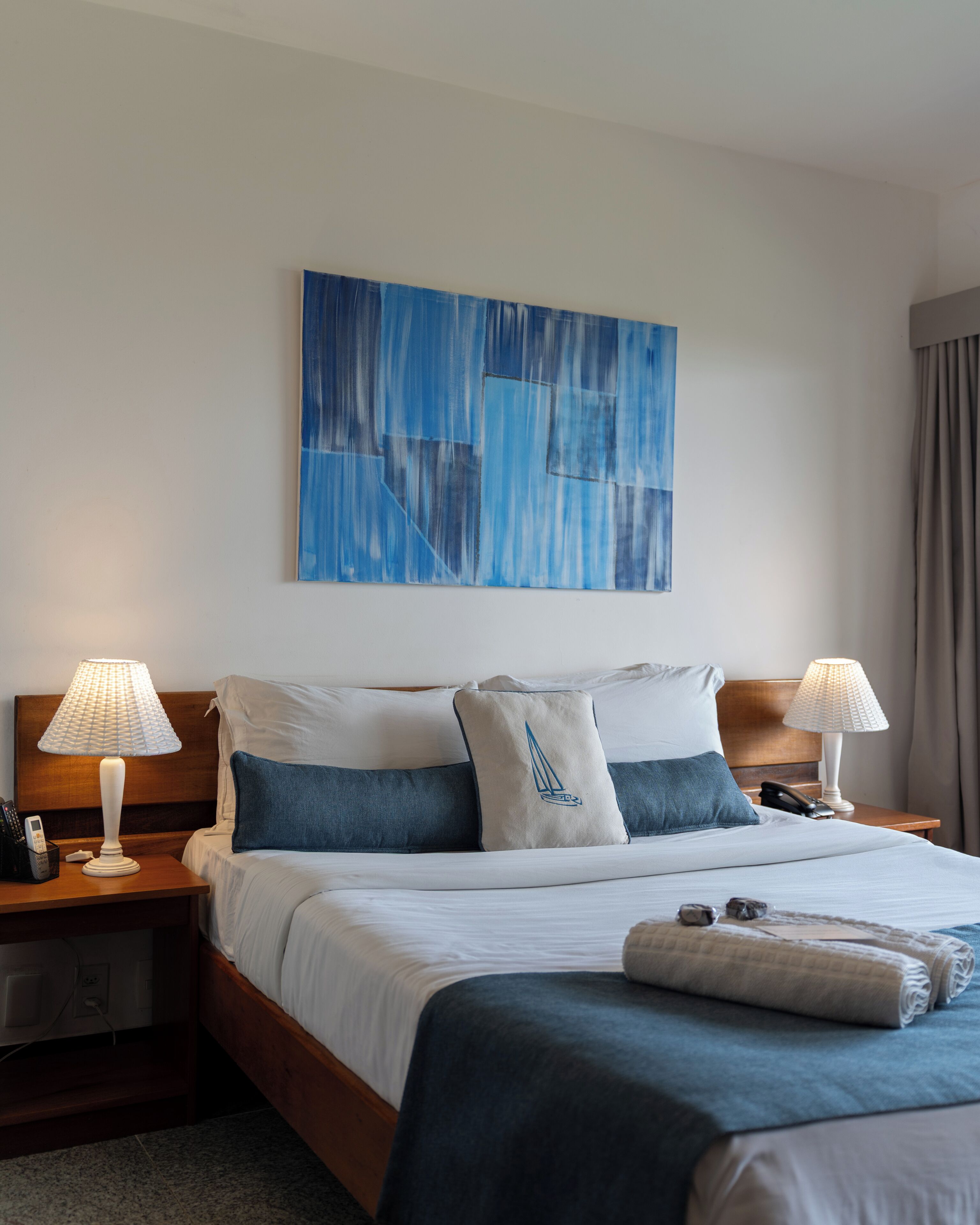 A hotel room with a bed, two bedside tables with lamps, a painting on the wall, and a window with curtains.