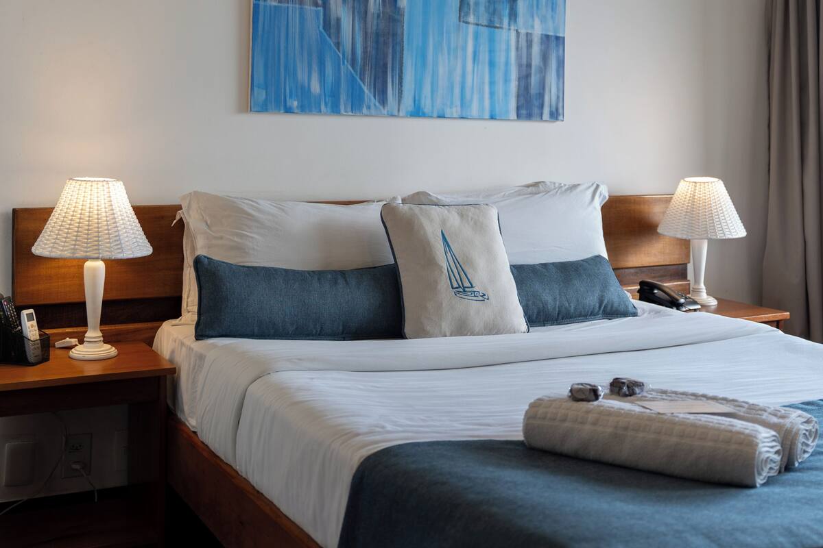 Una habitación de hotel con una cama, dos mesitas de noche con lámparas, un cuadro en la pared y una ventana con cortinas.