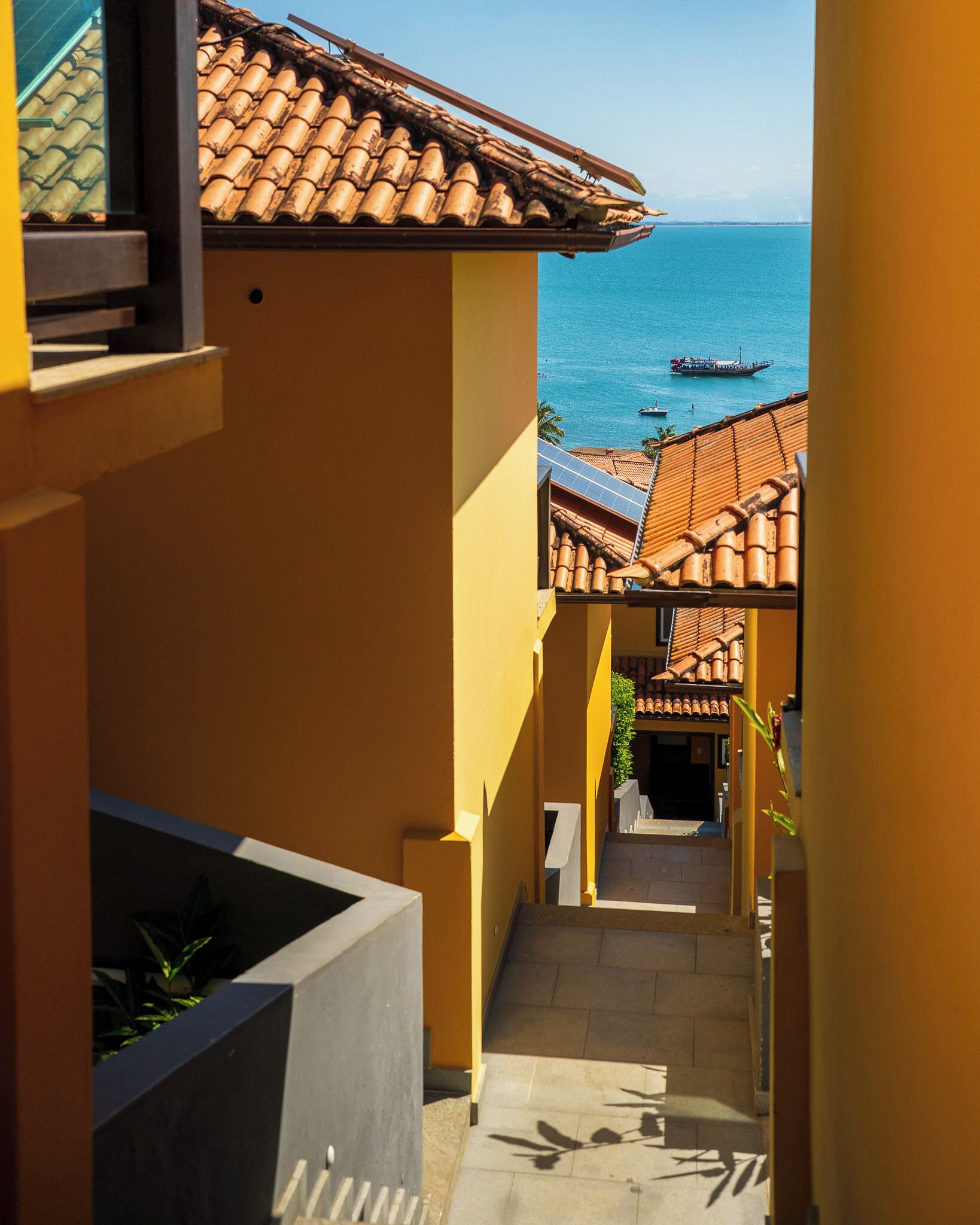 A narrow corridor with a view of the sea and boats, leading to a building with a tiled roof.
