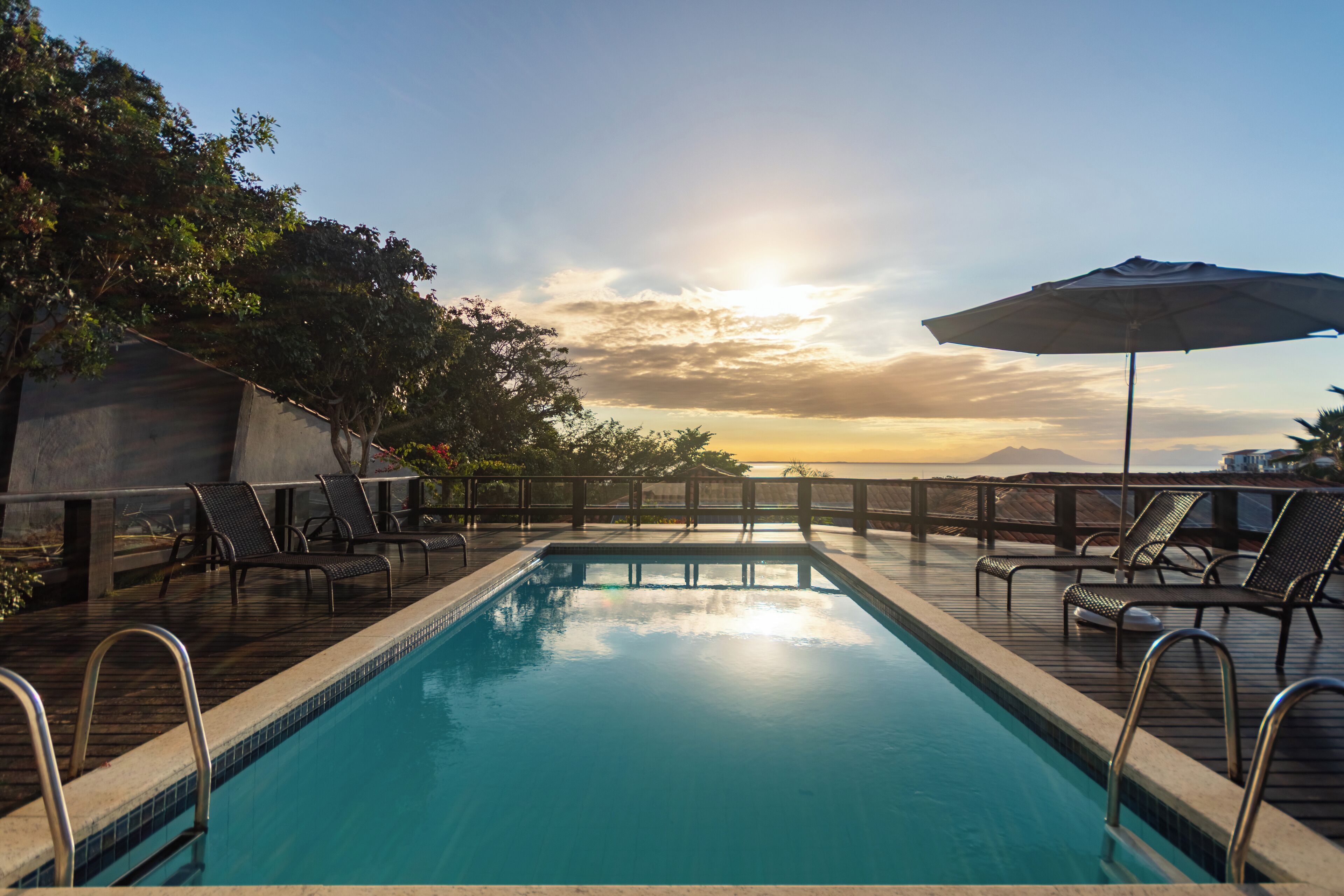 Una piscina con sillas de descanso y una sombrilla, rodeada por una terraza de madera y árboles, con un atardecer de fondo.