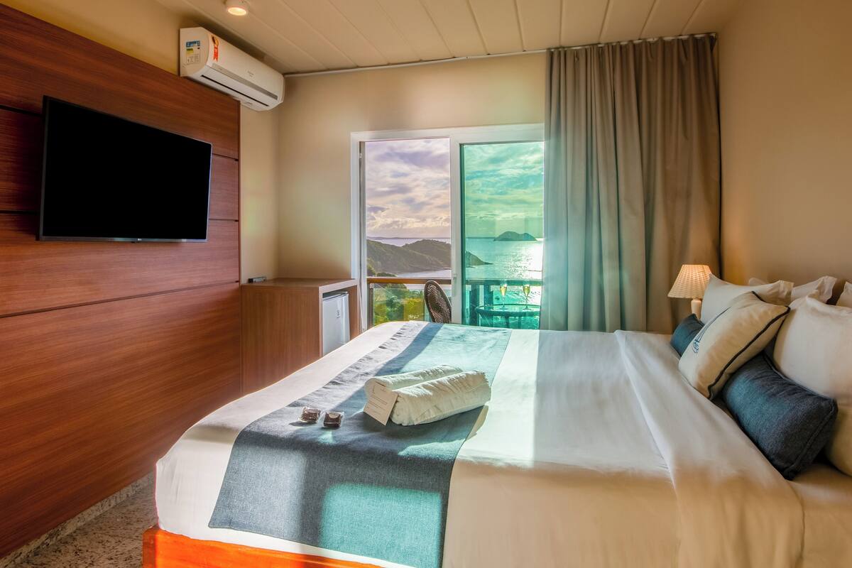 Habitación de hotel con una cama, un televisor de pantalla plana en la pared, una ventana con vista al mar y un equipo de aire acondicionado.