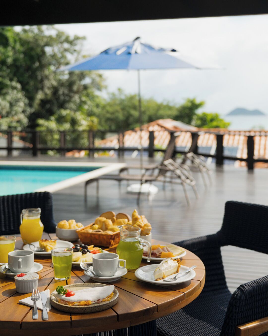 A breakfast table with various food items and drinks, overlooking a pool and greenery.
