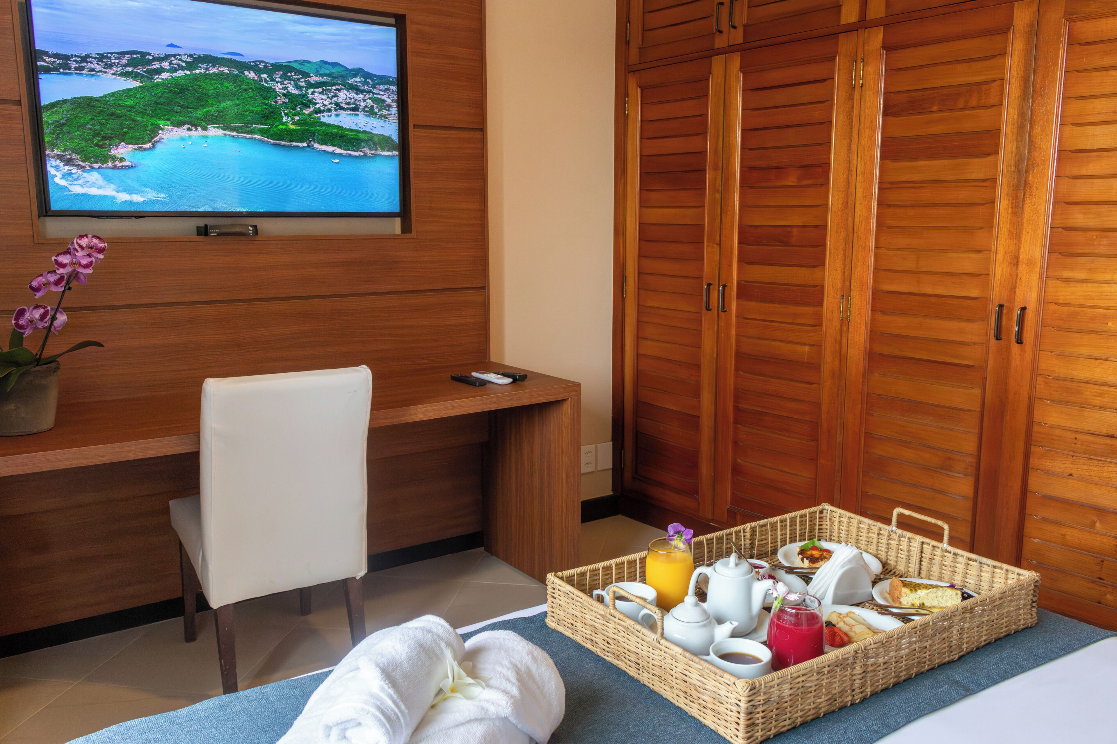 Habitación de hotel con un escritorio de madera, un televisor que muestra una vista panorámica, una bandeja de mimbre con alimentos para el desayuno y una cama con una toalla.