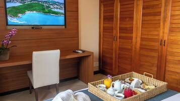 Habitación de hotel con un escritorio de madera, un televisor que muestra una vista panorámica, una bandeja de mimbre con alimentos para el desayuno y una cama con una toalla.