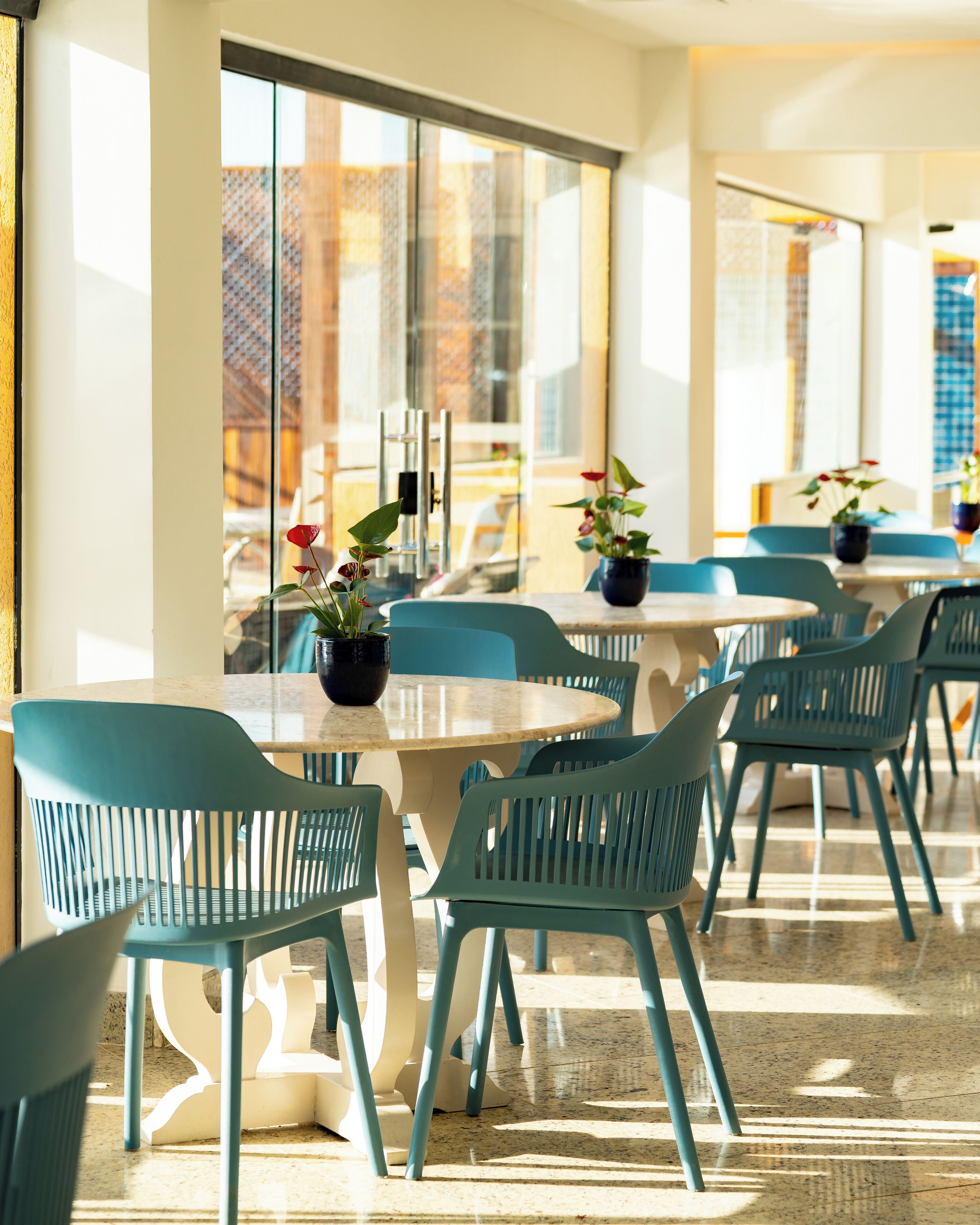 Een moderne eetruimte met ronde tafels en teal gekleurde stoelen, grote ramen met uitzicht op de stad en potplanten op de tafels.